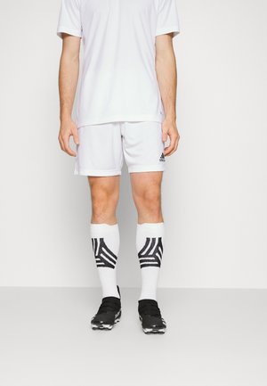 Person iført en hvid sportstøj med Adidas-shorts, hvide sokker med sorte mønstre og sorte fodboldstøvler, der står mod en hvid baggrund.
