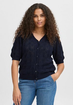 Cardigan tricoté bleu marine à manches courtes, avec un motif en dentelle texturée et des poignets et un ourlet côtelés. Petits boutons ronds sur le devant.
