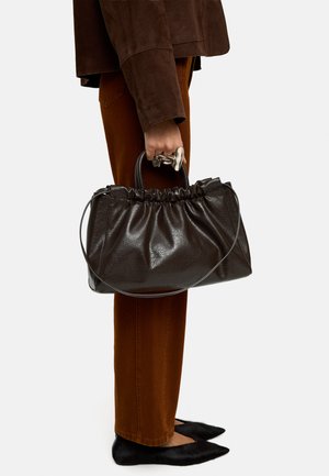 Osoba ubrana w brązowe spodnie i ciemnobrązową kurtkę, trzymająca dużą ciemnobrązową skórzaną torebkę z marszczonym wierzchem i cienkim paskiem na ramię.