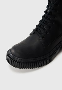 Černá kožená bota s kulatou špičkou, silnou černou gumovou podrážkou s hladkým textura, a černými tkaničkami. Hladký povrch s minimem detailů.