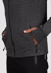 Dark gray zip-up jacket features a textured fabric, side pockets, and ribbed cuffs. The design is close-fitting with adjustable hardware.