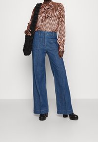 Bredde denimbukser i lyseblå farve med synlige sømme, parret med en mønstret bluse med flæsekrave. Sort håndtaske og kraftige hæle.