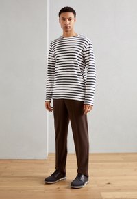 Striped long-sleeve shirt in black and white, brown trousers, and dark grey shoes with a textured finish, standing on a wooden floor.