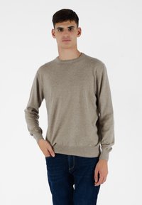 Jasno beżowy sweter z okrągłym dekoltem, długimi rękawami oraz prążkowanymi mankietami i dołem. Noszony z ciemnoniebieskimi dżinsami, na jednolitym białym tle.