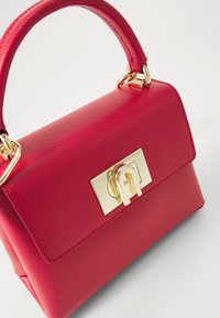 Sac à main en cuir rouge, de forme structurée, avec des accessoires dorés et un rabat plat à l'avant sécurisé par un fermoir. Surface texturée avec une petite poignée.