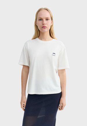 Blondynka w białej koszulce z małym nadrukiem filiżanki na piersi i czarnej spódnicy, stojąca na jednolitym tle.