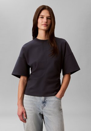 Vrouw met lang bruin haar draagt een loszittend donkergrijs T-shirt met korte mouwen en lichtblauwe spijkerbroek, staand met één hand in de zak op een grijze achtergrond.