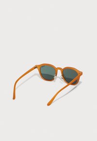 Gafas de sol de plástico naranja con lentes verdes, forma de ojo de gato, textura suave y brazos delgados. Los detalles notables incluyen herrajes de metal en las bisagras.