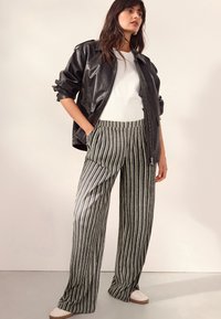 Giacca in ecopelle nera, t-shirt bianca, pantaloni a palazzo a righe in bianco e nero, e sneakers beige. Tessuto morbido, vestibilità casual.
