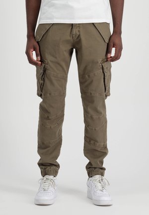 Mand iført brune cargobukser med klaplommer og hvide sneakers, der står mod en ensfarvet baggrund.