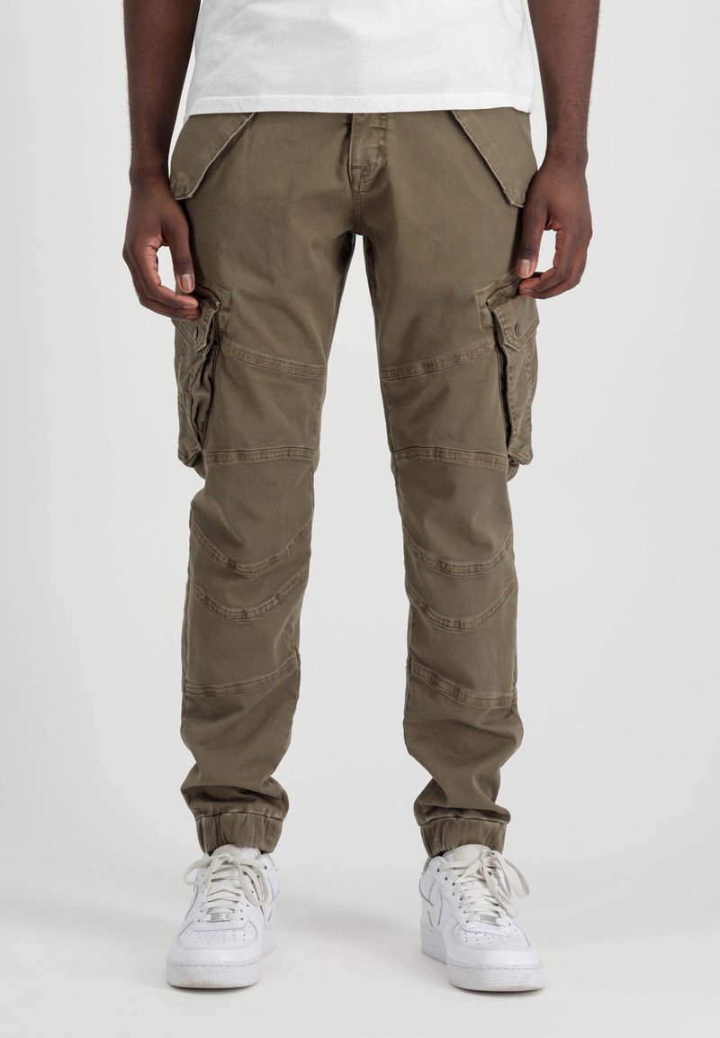 Homme portant un pantalon cargo marron avec poches à rabat et des baskets blanches, debout devant un fond uni.