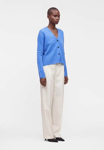 Femme debout portant un cardigan bleu boutonné, un pantalon blanc à jambes larges et des chaussures pointues noires sur un fond blanc uni.