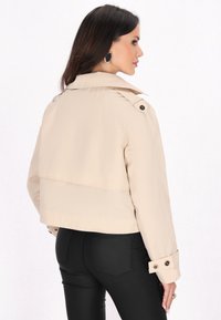 Beige cropped dzseki vállpántokkal, dupla gombos záródással és gombbal rögzített mandzsettákkal; fekete, szűkített nadrággal párosítva. Sima textúra.