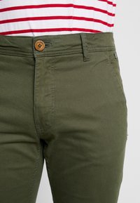 Olivgrüne Hose aus weicher Baumwolle, mit vorderem Knopfverschluss, flacher Vorderseite und dezenten Nahtdetails entlang der Naht.