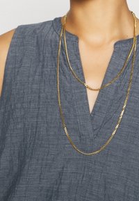 Deux chaînes en or de longueurs différentes se drapent sur un haut texturé bleu-gris à encolure en V et fines rayures verticales.