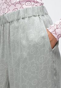 Roka gaismēpelēs pelēkās biksēs ar baltu ziedu rakstu, valkājot rozā ar ziedu rakstu garām piedurknēm topu.