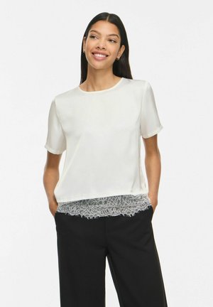 Femme aux longs cheveux foncés portant un chemisier blanc à manches courtes avec bordure en dentelle et un pantalon noir, souriante et debout les mains dans les poches.