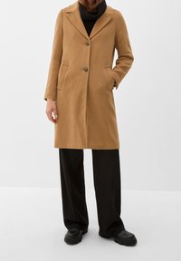 Beżowy wełniany płaszcz z wyłogami, dwoma przednimi kieszeniami i zapięciem na guziki, zestawiony z czarnymi spodniami o szerokich nogawkach i masywnymi czarnymi botkami.