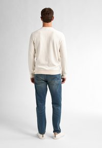 Heren lichtbeige langemouwen shirt met ribbelstructuur, gecombineerd met donkerblauwe jeans en witte sneakers, van achteraf bekeken.