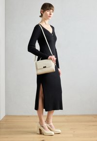 Robe noire côtelée avec des manches longues et une fente sur le côté. Le modèle porte un petit sac crème avec une surface texturée et des accessoires en or. Talons beiges.