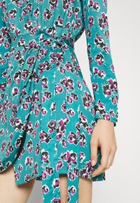Türkisfarbenes Blumenkleid mit schwarzen und magentafarbenen Rosen, langen Ärmeln, einer gebundenen Taille und Faltenrockdetails. Glatte Stofftextur.