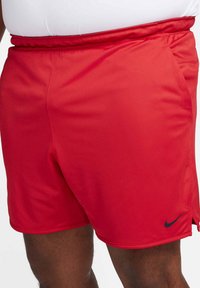 Uomo che indossa pantaloncini atletici rosso vivo con il logo Nike nero sulla gamba sinistra e vita elastica, mostrato dalla vita fino a metà coscia.