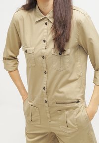 Tuta utilitaria beige con maniche lunghe, chiusura con bottoni e due tasche sul petto. Presenta una tasca laterale con zip e una vestibilità comoda.