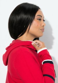 Czerwona bluza z kapturem z białymi i granatowymi paskami na mankietach, teksturowana tkanina oraz kaptur ze sznurkiem. Modelka przedstawiona na zdjęciu profilowym.