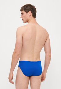 Blå svømmebukser med en glat tekstur og elastisk talje, set fra bagsiden, med en tætsiddende pasform og minimal dækning.