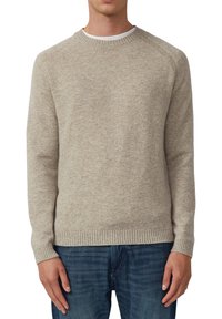 Mężczyzna w beżowym swetrze typu crewneck i niebieskich dżinsach, stojący na białym tle z rękami luźno ułożonymi po bokach.