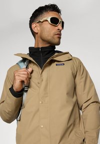 Brun vanntett jakke med trykknapper, glidelåslomme og justerbar hette, lagd over en svart zip-up topp, båret med kremfargede solbriller.