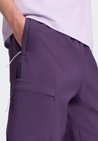 Paarse cargo-broeken met een gladde textuur, voorzien van zijzakken en een trekkoord voor aanpassing. Gedragen met een lichtpaarse top.