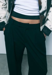 Črne plisirane hlače z detajlom v obliki mašne na pasu, v kombinaciji z belo top majico in vzorčastim krznom, ki ima rjave in sive kontraste.