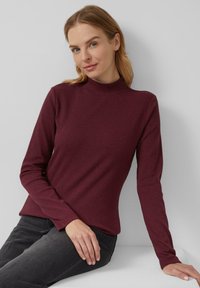 Vrouw met licht haar in een getailleerde, lange mouwen, kastanjebruine opstaande kraag top en zwarte jeans, zittend tegen een effen lichtgrijze achtergrond.