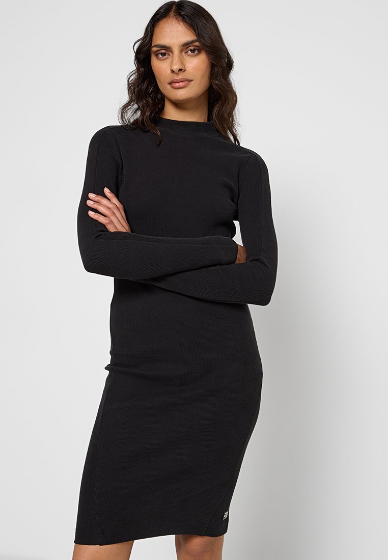 Femme avec des cheveux sombres et ondulés portant une robe noire ajustée à manches longues, longueur genou, se tenant les bras croisés devant un fond uni.