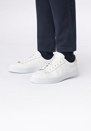 Weiße Leder Sneaker mit glatter Textur, runder Zehenpartie und perforierten Akzenten. Verfügt über eine flache weiße Sohle und einen braunen Fersendetail, getragen mit dunklen Hosen.