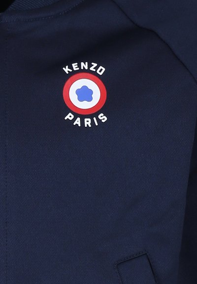 Veste marine ornée d'un logo rond avec une fleur bleue et le texte "KENZO PARIS". Tissu lisse avec des détails brodés et un design moderne.