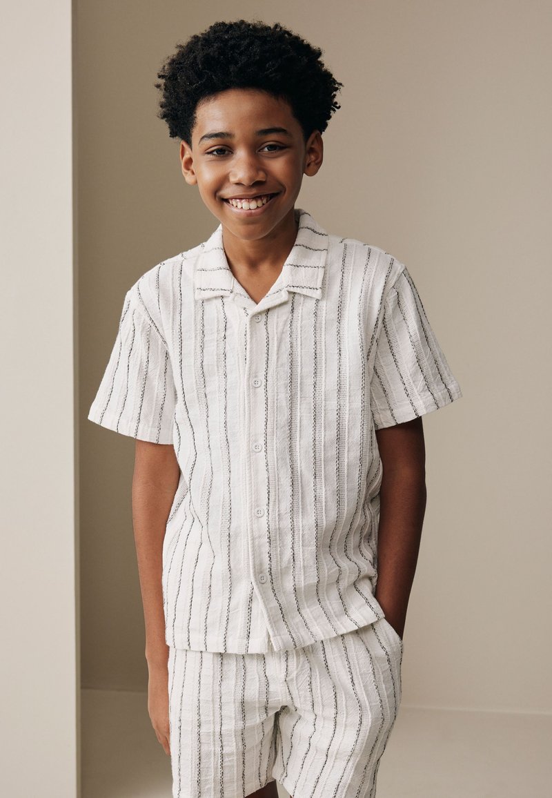 Niño sonriendo con las manos en los bolsillos, vestido con camisa blanca de manga corta con botones y pantalones cortos a juego con finas rayas verticales negras.