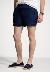 Navy blue linen shorts with a drawstring waist, side pockets, and a small red emblem. Paired with navy espadrilles featuring a jute sole.