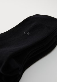 Schwarze Socken aus einem glatten Stoff, ordentlich gefaltet, mit einem dezenten grauen "ck"-Logo auf einer Seite.