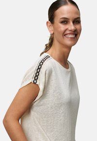 Lys beige t-shirt lavet af struktureret stof med sort blomsterbroderi langs skulderen, med korte ærmer og en afslappet pasform.