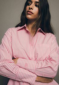 Różowa w paski koszula na guziki z kołnierzykiem i kieszonką. Posiada długie rękawy, zapinane na guziki mankiety oraz subtelne hafty na rękawie.