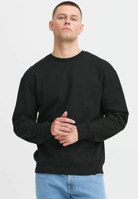 Sudadera negra de manga larga, cuello redondo y puños acanalados. Tejido suave, corte relajado, exhibida en un modelo con un fondo neutro.