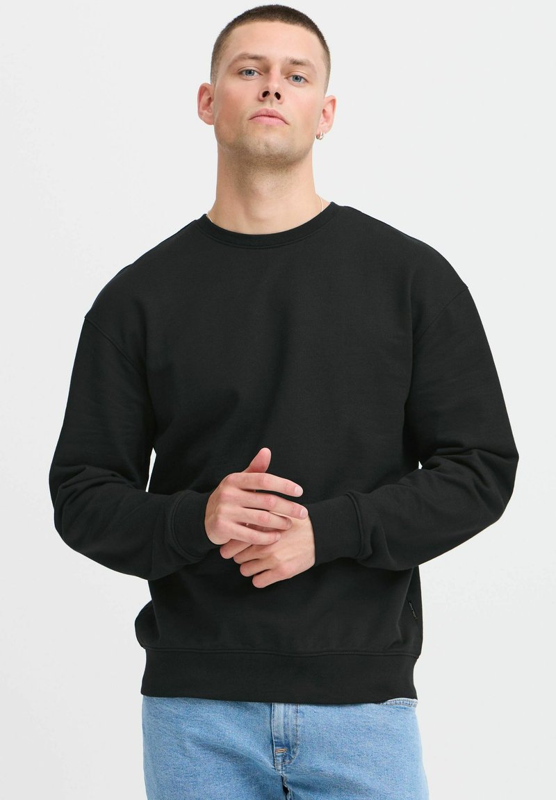 Sudadera negra de manga larga, cuello redondo y puños acanalados. Tejido suave, corte relajado, exhibida en un modelo con un fondo neutro.