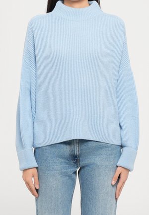 Pull en maille côtelée bleu clair avec col montant et poignets roulés, porté avec un jean bleu délavé sur une personne debout.