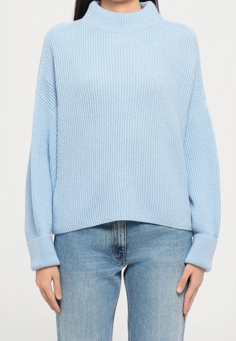 Pull en maille côtelée bleu clair avec col montant et poignets roulés, porté avec un jean bleu délavé sur une personne debout.