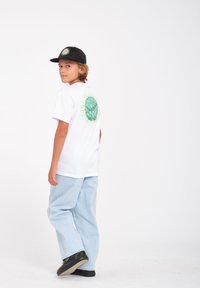 T-shirt blanche avec un graphique vert au dos, associée à un jean baggy bleu clair et une casquette noire présentant un design vert similaire.