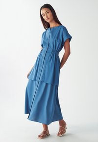 Modra obleka s kratkimi rokavi, elastičnim pasom, večplastnim robom in gladko teksturo, v kombinaciji s sandali brez pete.