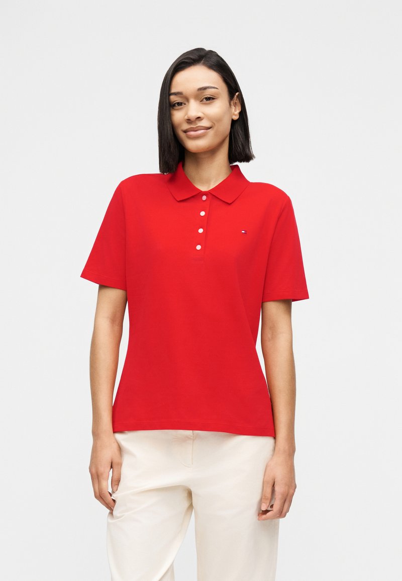 Γυναίκα με ίσια σκούρα μαλλιά, ντυμένη με κόκκινο πόλο κοντομάνικο και κρεμ παντελόνι, στέκεται μπροστά σε απλό φωτεινό φόντο.