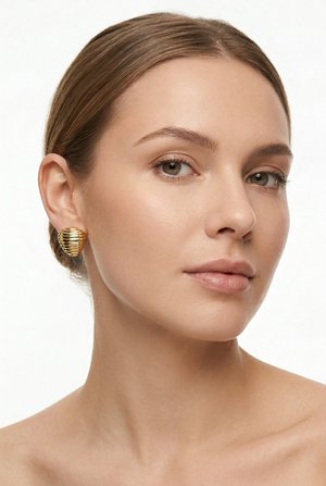 Vaaleaihoinen nainen, jolla on ruskeat hiukset vedettynä taakse, pukeutuneena suuriin kultaisiin uurteisiin simpukkakorvakoruihin, katsoo hieman sivulle.